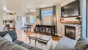 A spacious living room with a large TV, a fireplace, and a kitchen in the background.