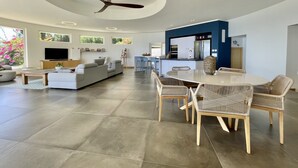 Een moderne woonkamer met een grote bank, een eettafel met rieten stoelen, een plafondventilator en een keuken met een blauwe accentmuur.