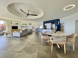 A modern living room with a large sofa, a dining table with wicker chairs, a ceiling fan, and a kitchen area with a blue accent wall. - Villa Menemene | Punaauia • Private Pool • Moorea View • 4 Bedrooms (PUNAAUIA)