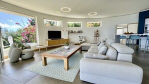 Een moderne woonkamer met een bank, salontafel en televisie. Er is een balkon met bloemen en een zichtbare keukenhoek.
