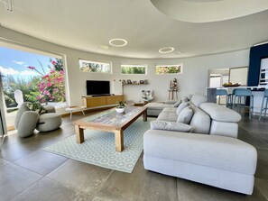 A modern living room with a sofa, coffee table, and TV. There is a balcony with flowers and a kitchen area visible. - Villa Menemene | Punaauia • Private Pool • Moorea View • 4 Bedrooms (PUNAAUIA)