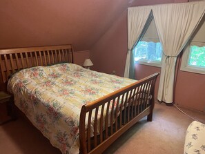 Un dormitorio con cama de madera, una mesita de noche, una lámpara, una ventana con cortinas y vistas al exterior con vegetación.