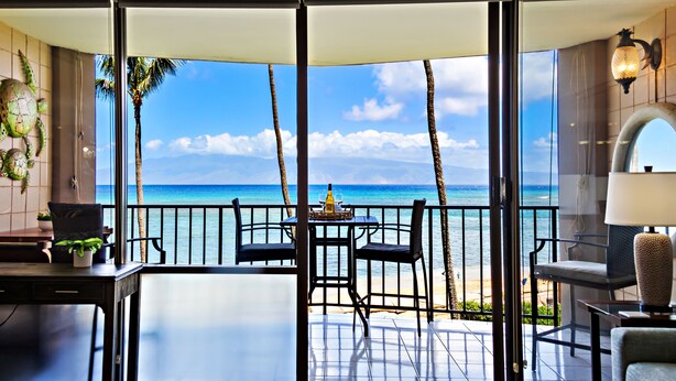 Un balcon avec vue sur l’océan, comprenant une table et des chaises, des palmiers et une lampe décorative.