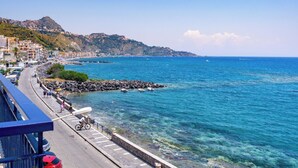 A coastal road with a stone railing, clear blue sea, and buildings on a hillside.