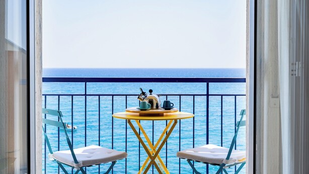 A balcony with a view of the sea, a small round table with a kettle and cups, and two chairs.