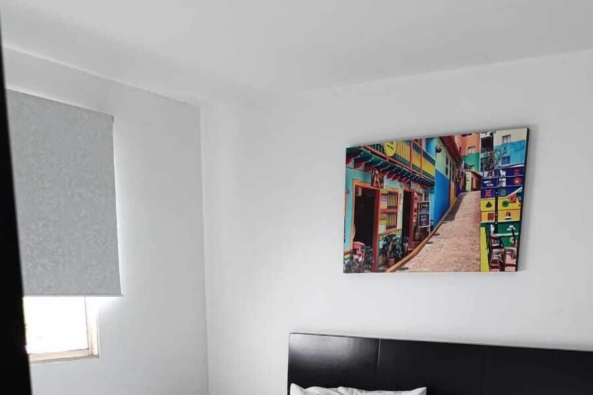 Un dormitorio moderno con cama, ropa de cama blanca, una manta azul, cabecera negra, mesita de noche, un cuadro colgado en la pared y una ventana con persianas.