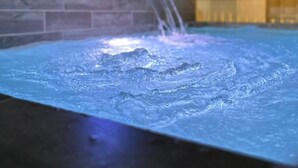 Una piscina con una cascada, rodeada por un muro de piedra y un edificio con techo azul al fondo.