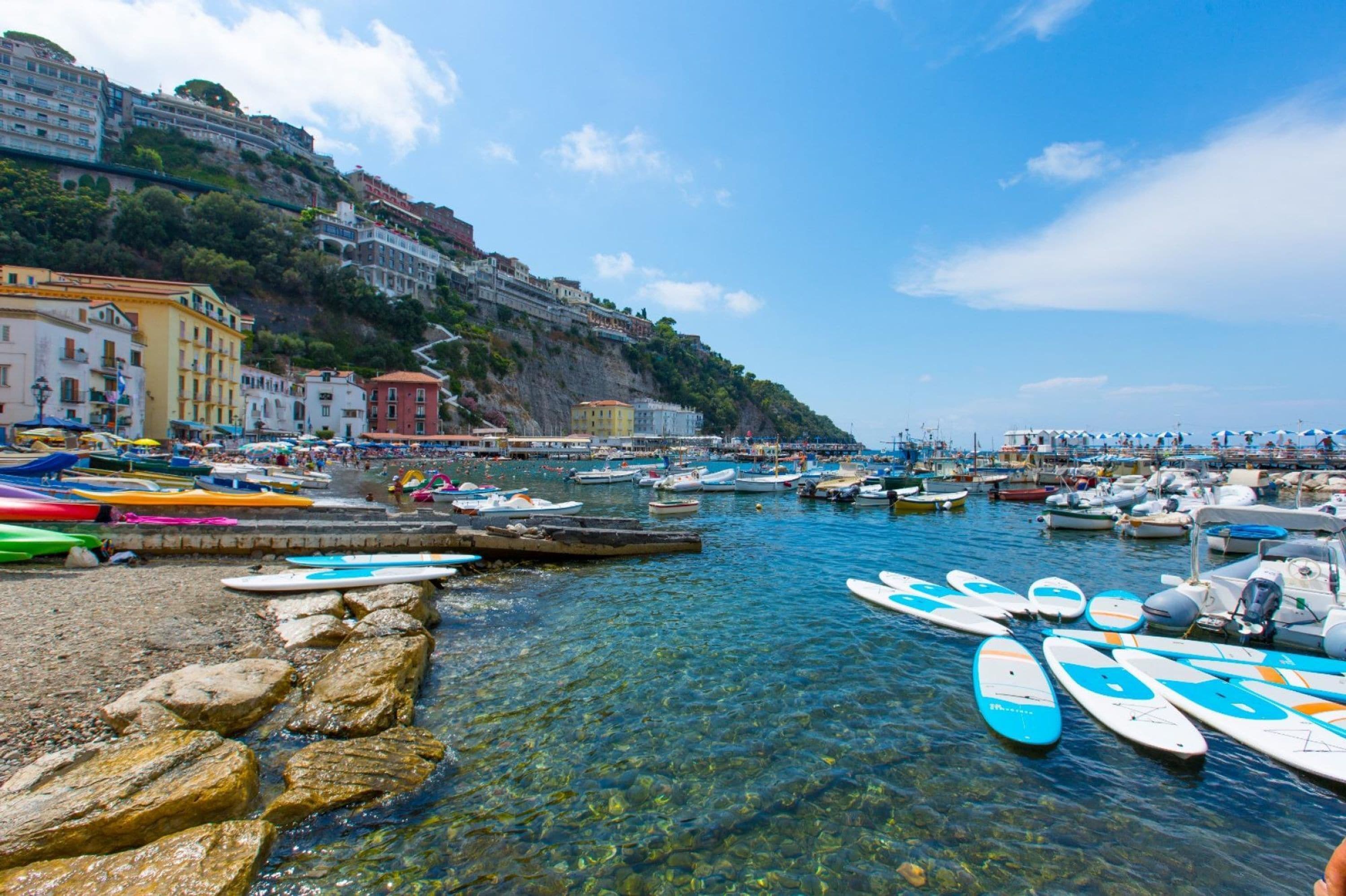 A coastal marina with boats and surfboards, buildings along the cliff, and clear blue water.