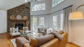 A living room with a stone fireplace, a leather sofa, a glass coffee table, and a large window with curtains. - Bryson City Mtn Retreat w/ Hot Tub (Bryson City)