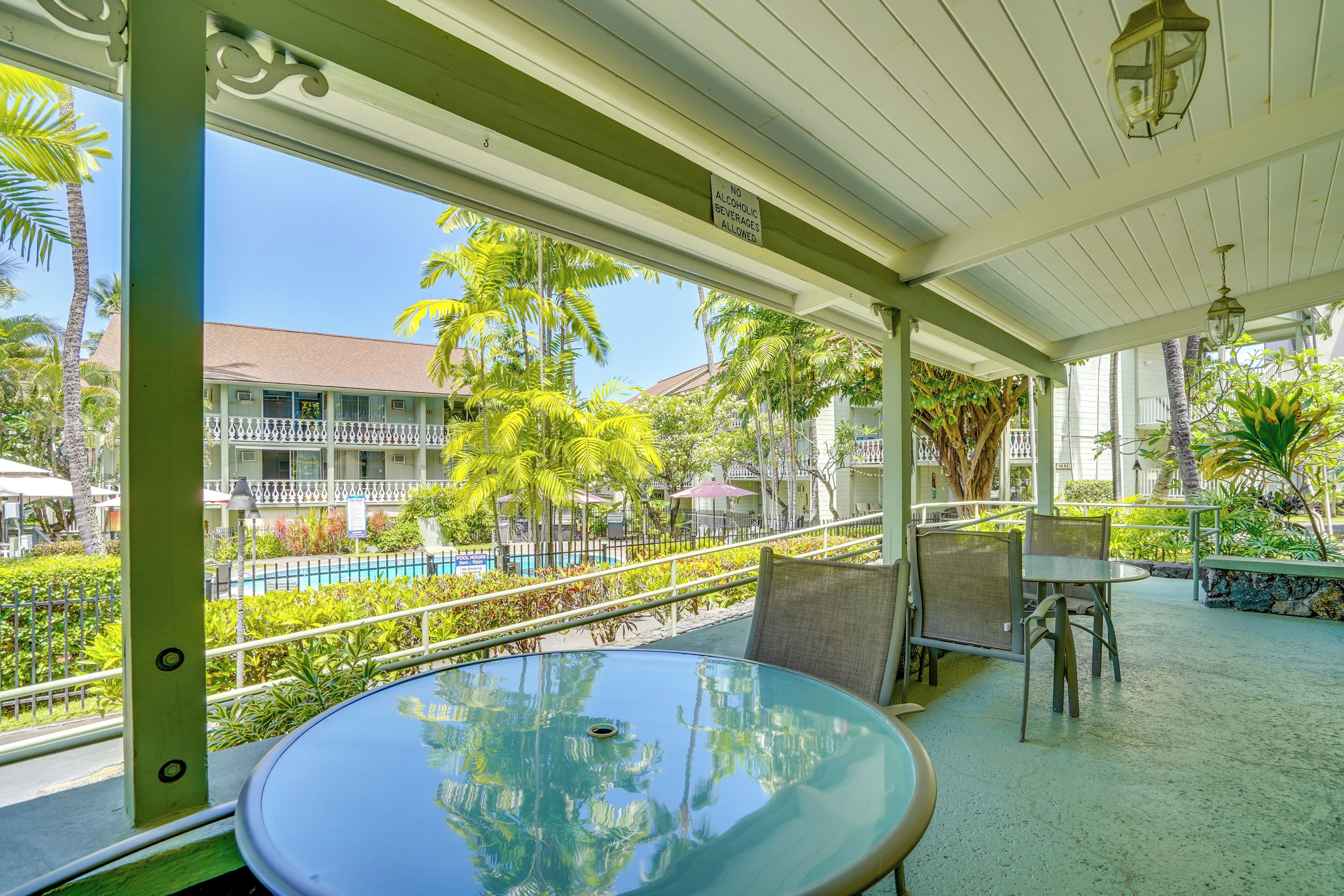 A covered outdoor area with a glass table and chairs, a pool in the background, and a sign prohibiting alcohol and beverages.