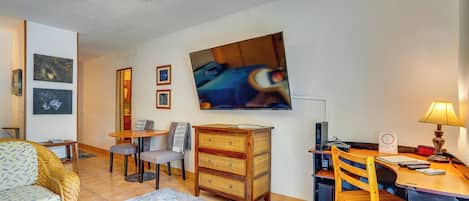 A living room with a ceiling fan, a television mounted on the wall, a dining table with chairs, a wicker sofa, and a desk with a lamp.