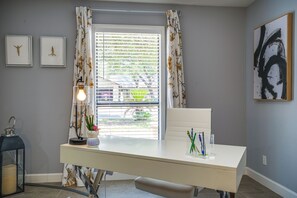 Um quarto com uma mesa, janela com persianas, um candeeiro e quadros nas paredes.