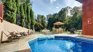 A swimming pool with lounge chairs and an umbrella, surrounded by trees and a wooden fence.