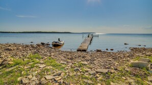 A rocky shoreline with a dock extending into calm water, a boat moored at the dock's end, and a clear sky above. - Prairie-style Home on Garden Bay w/ Deck + Hot Tub (Garden)