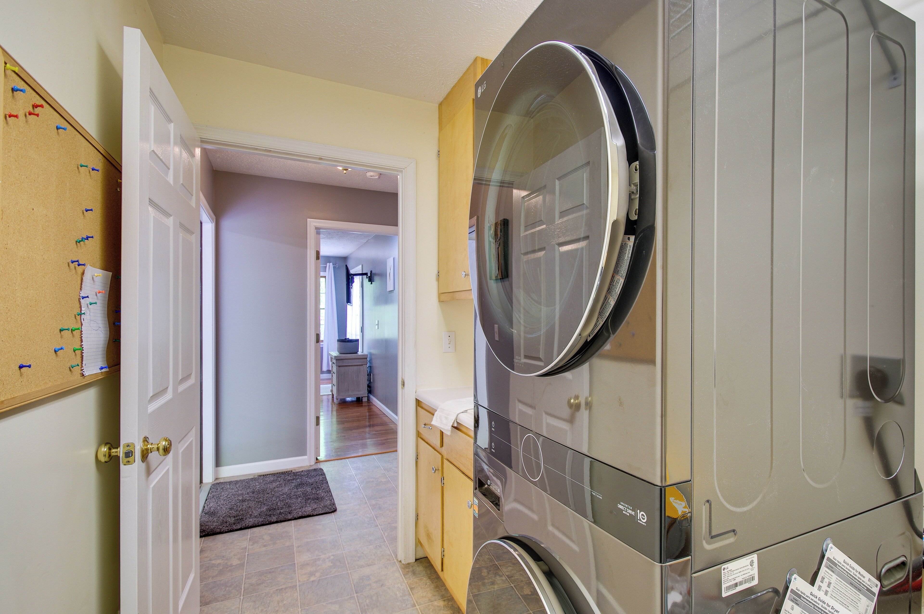 A laundry room with a large front-loading washer and dryer, a corkboard with pins and notes, and a door leading to a hallway.