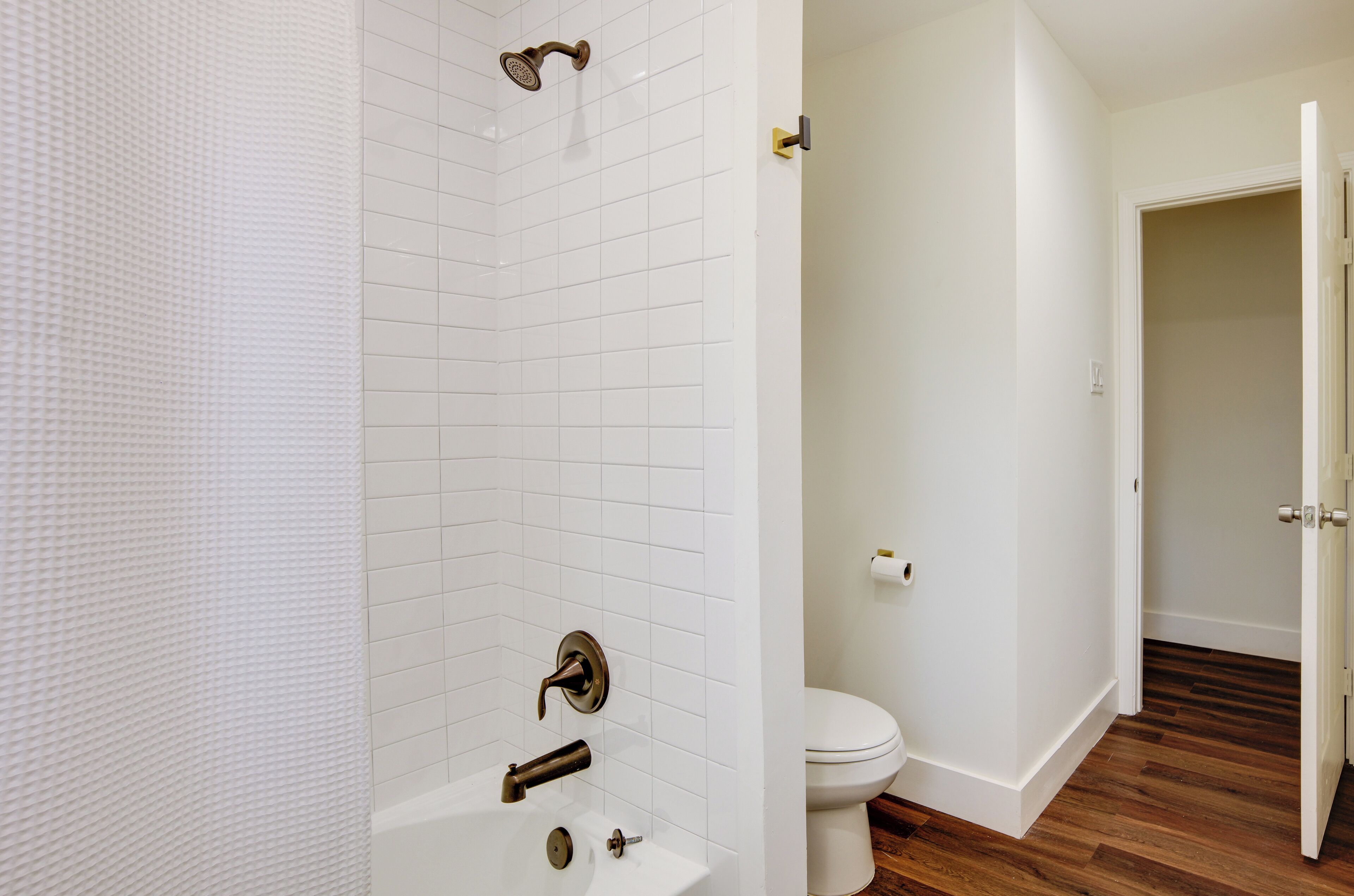 Un baño con paredes de azulejos blancos, un inodoro blanco y piso de madera.