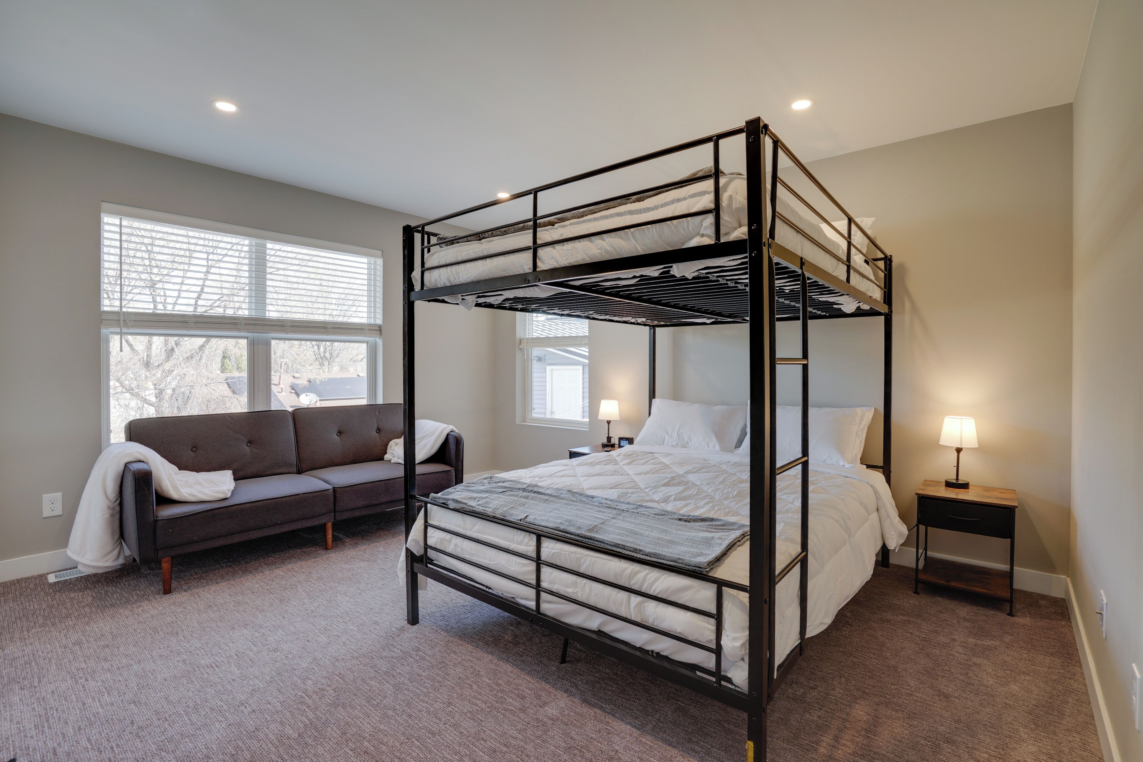 A bunk bed room with a sofa, bedside tables, and a window with blinds.