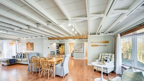 A spacious living area with a wooden dining table, chairs, and a sofa. There are wooden beams on the ceiling, and a large window on the right side. - Grand Junction Lake Home: 13 Mi to South Haven! (Grand Junction)