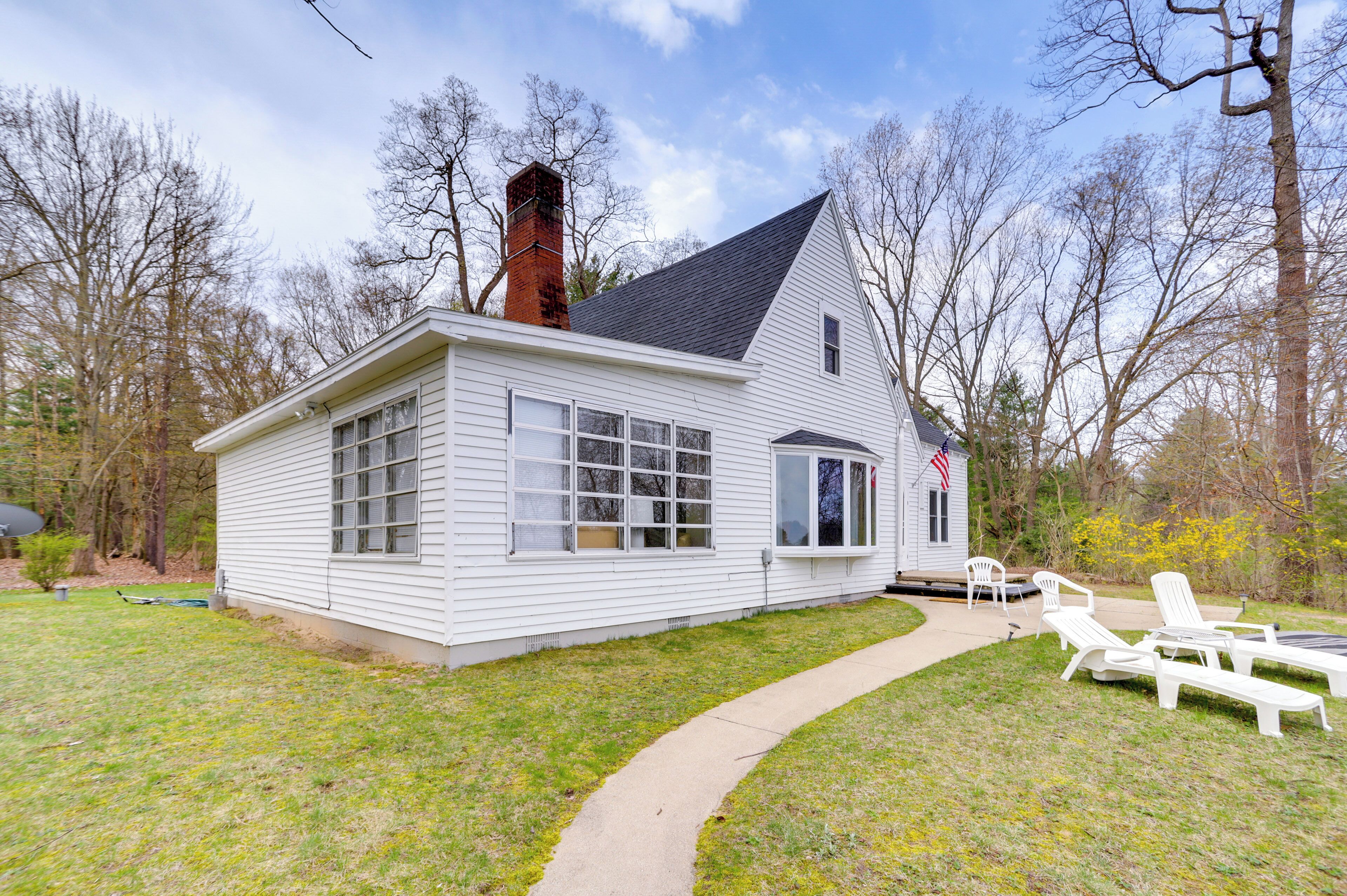 A white single-story house with a chimney, large windows, and a small porch with lawn chairs.
