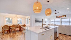 A modern kitchen with a white island, dining area with wicker chairs, and large windows offering a view. - East Jordan Vacation Rental on Lake Charlevoix! (East Jordan)
