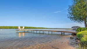 A wooden dock extending into a calm lake with clear water, surrounded by greenery and a clear sky. - East Jordan Vacation Rental on Lake Charlevoix! (East Jordan)