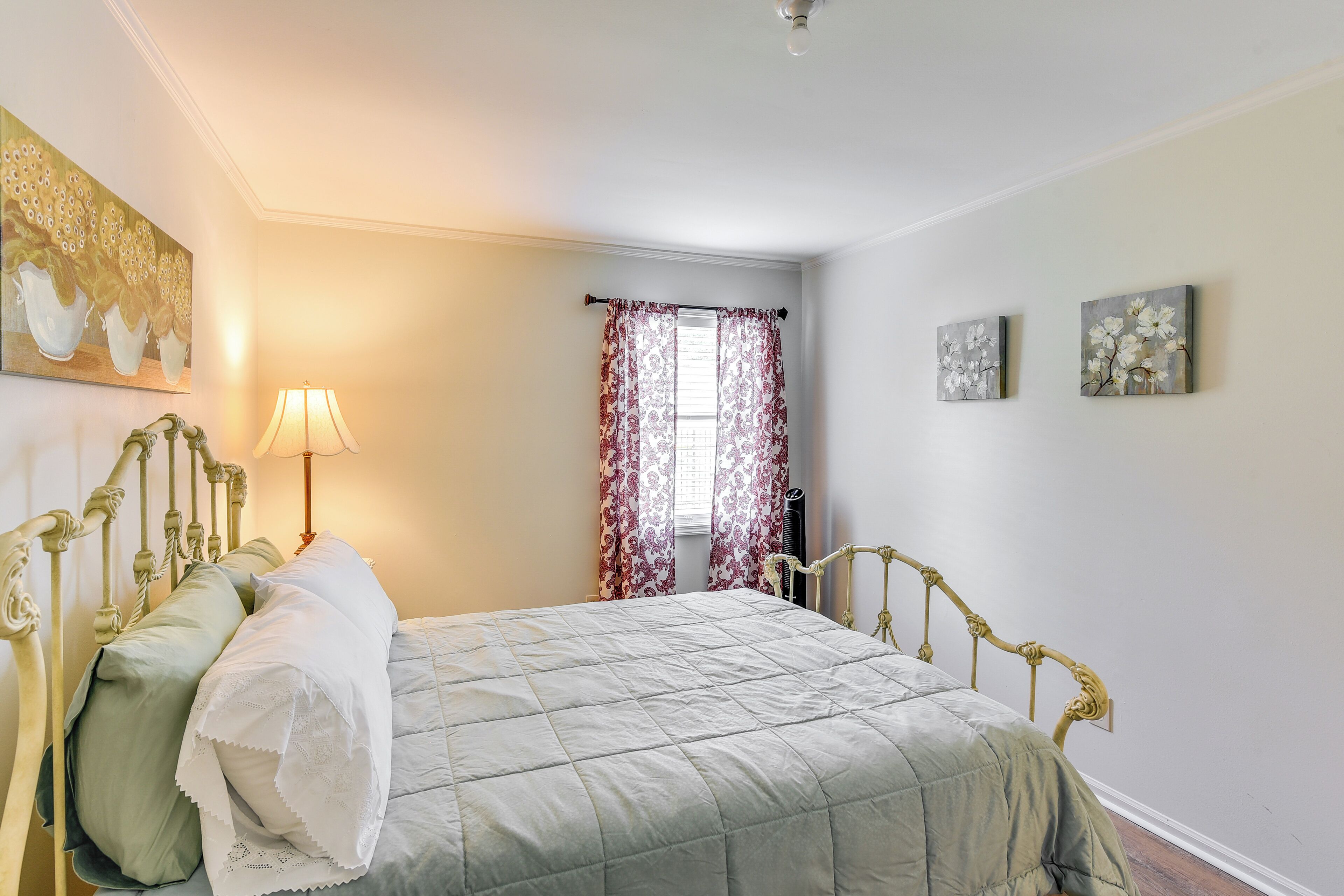 Een slaapkamer met een bed, een lamp, een raam met gordijnen en twee schilderijen aan de muur.