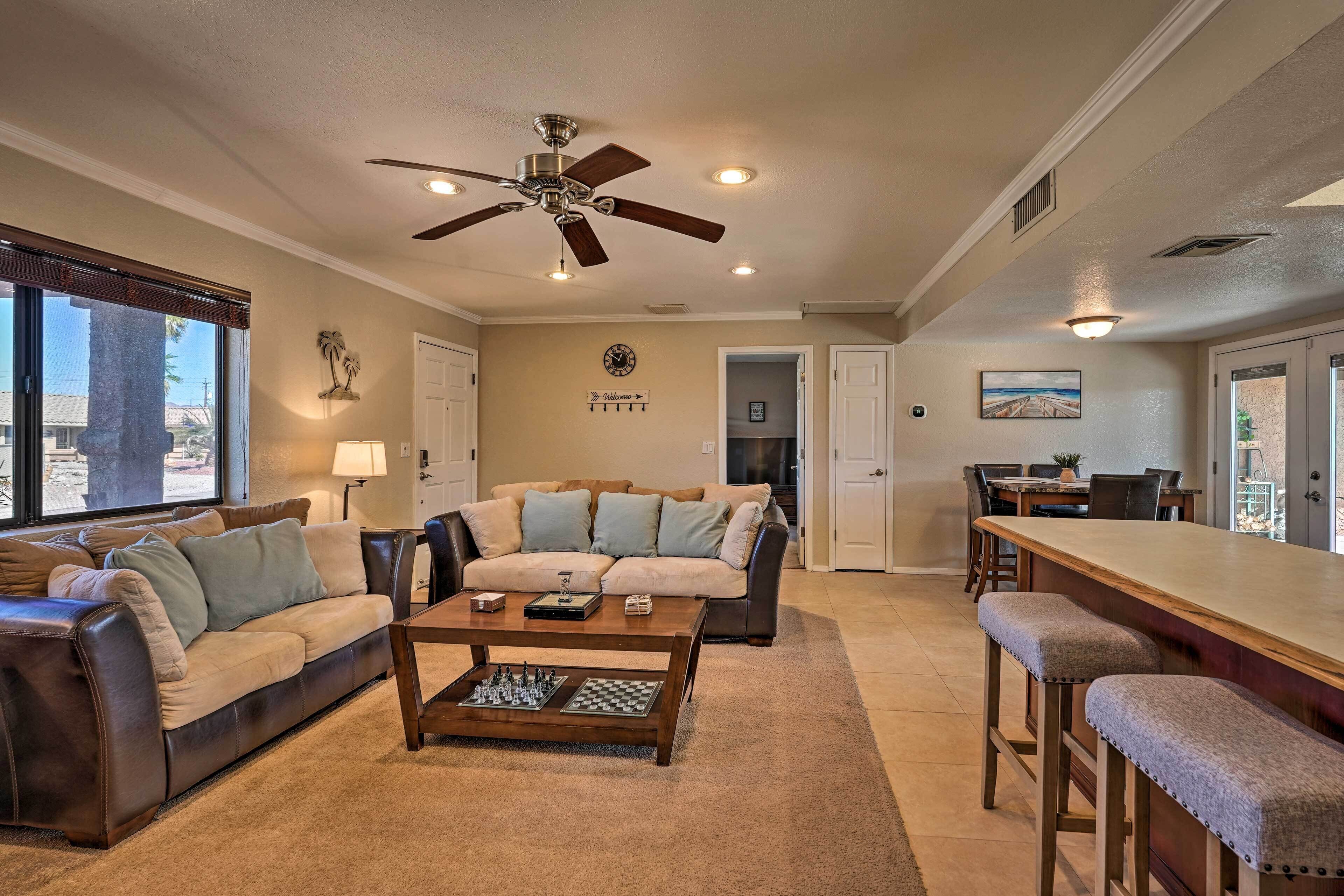 A spacious living room with a large sofa, a coffee table with chess pieces, a ceiling fan, and a view of the outside.