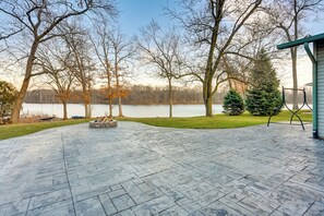 A patio with a stone floor, a fire pit, and a view of a lake with trees and a building in the background.
