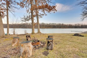A lakeside campsite with four wooden stumps arranged in a circle, a fire pit, and a view of the lake and trees.