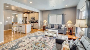 A modern living room with a grey sofa, a coffee table, and a patterned rug. There is a kitchen area with wooden cabinets and a refrigerator.