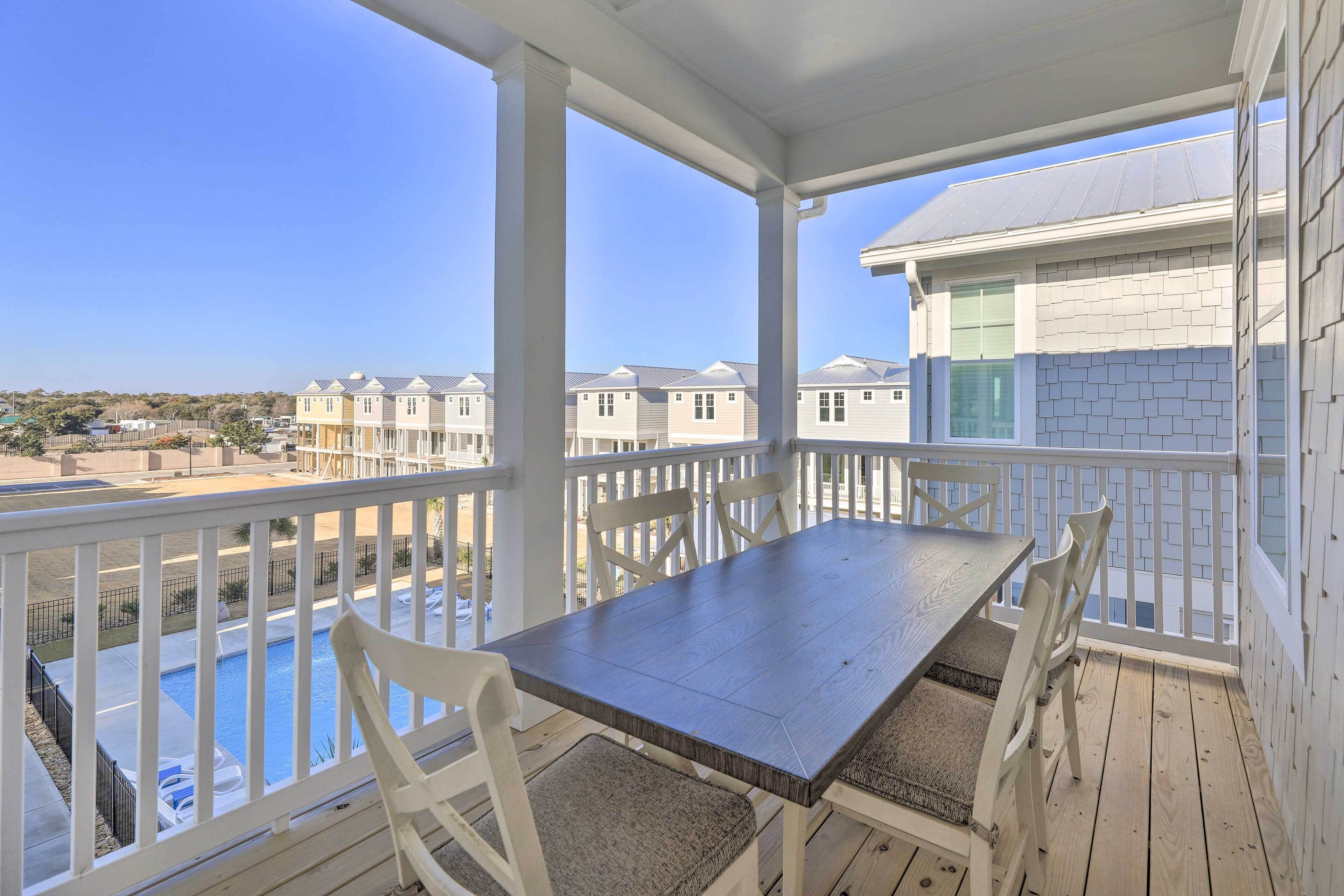 Varanda coberta com mesa de madeira e cadeiras brancas, com vista para uma piscina e casas vizinhas.