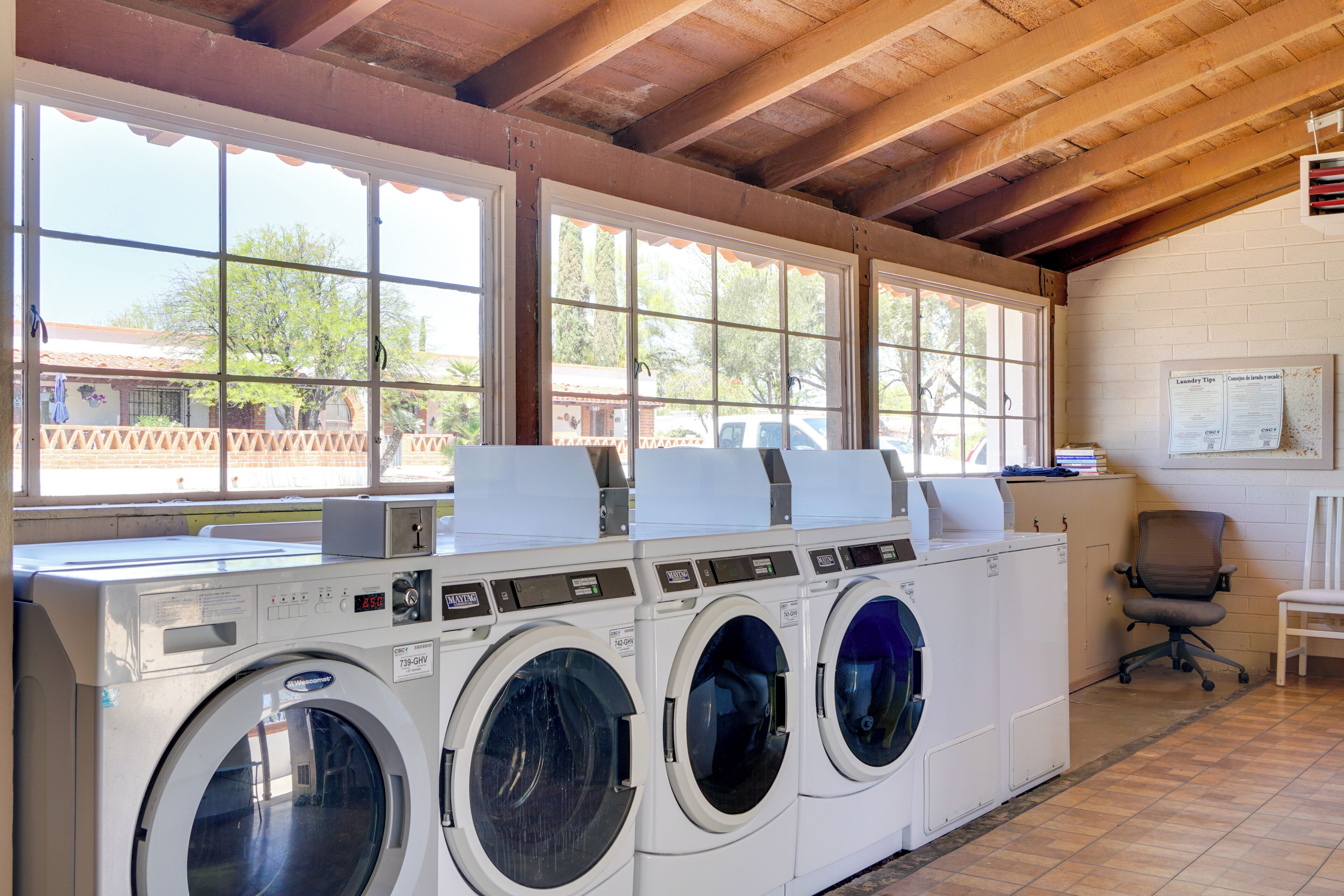A laundry facility with multiple washing machines and dryers, large windows, and a wooden ceiling.