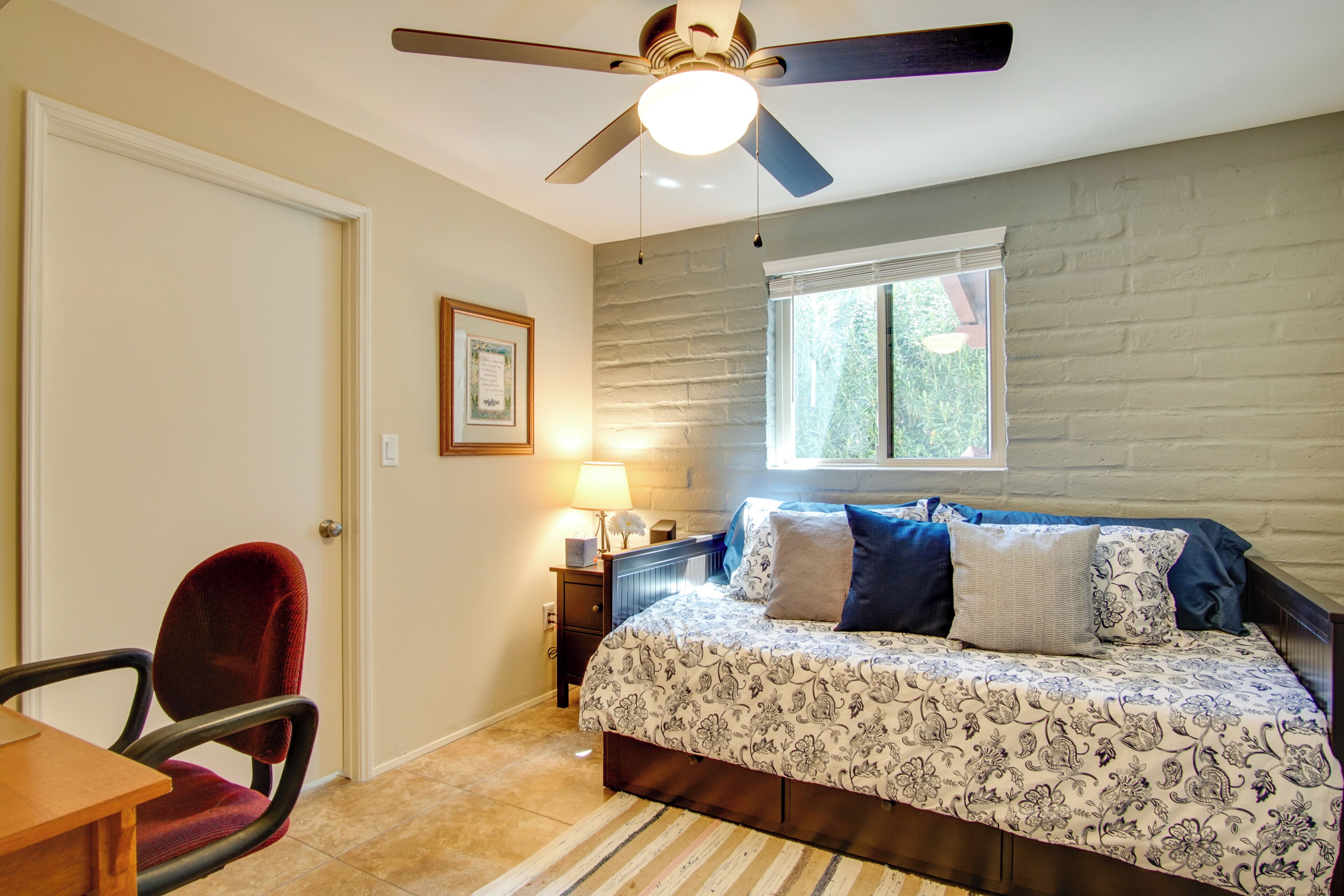 A bedroom with a bed, a chair, a desk, a ceiling fan, a window, and a lamp.