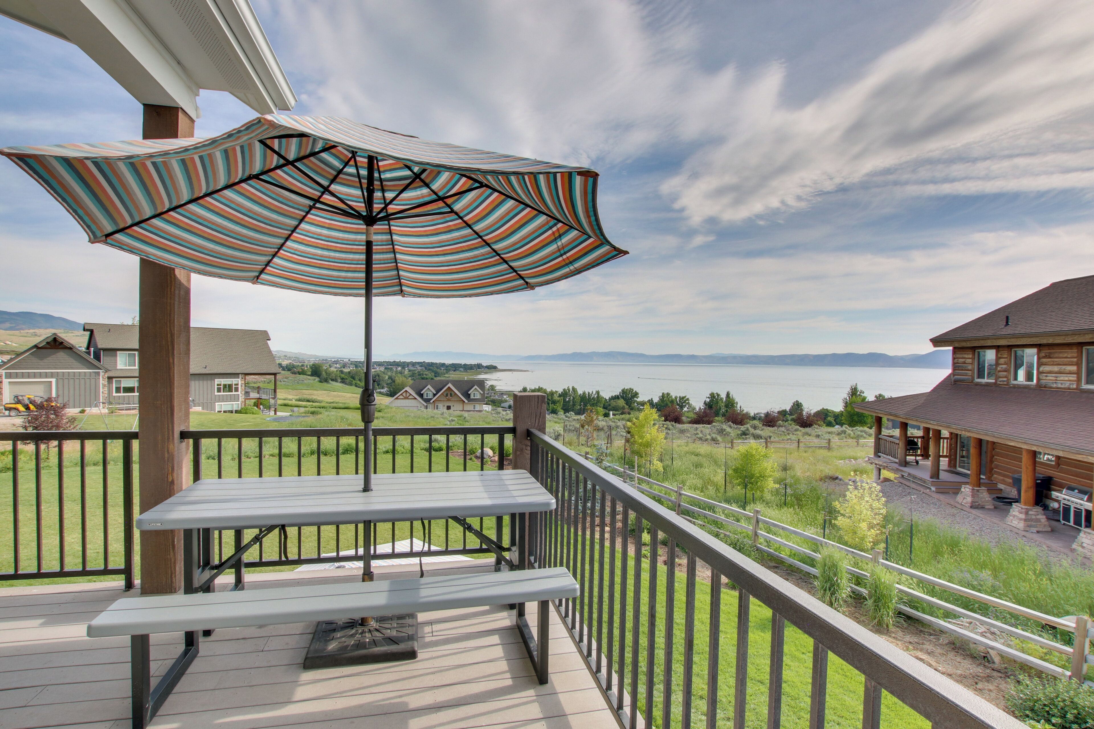 Une terrasse en bois avec une table de pique-nique et un parasol à rayures, d’où l’on a vue sur un lac et sur des maisons.