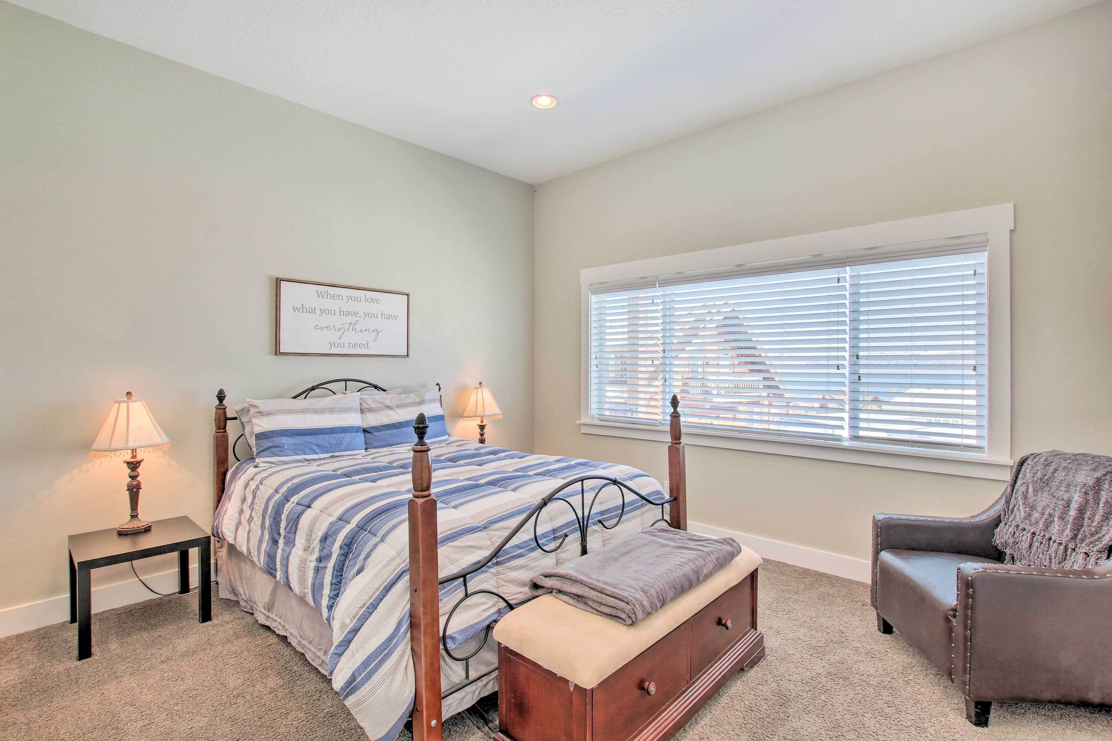 A bedroom with a bed, bedside lamps, a window with blinds, a chair, and a framed quote on the wall.
