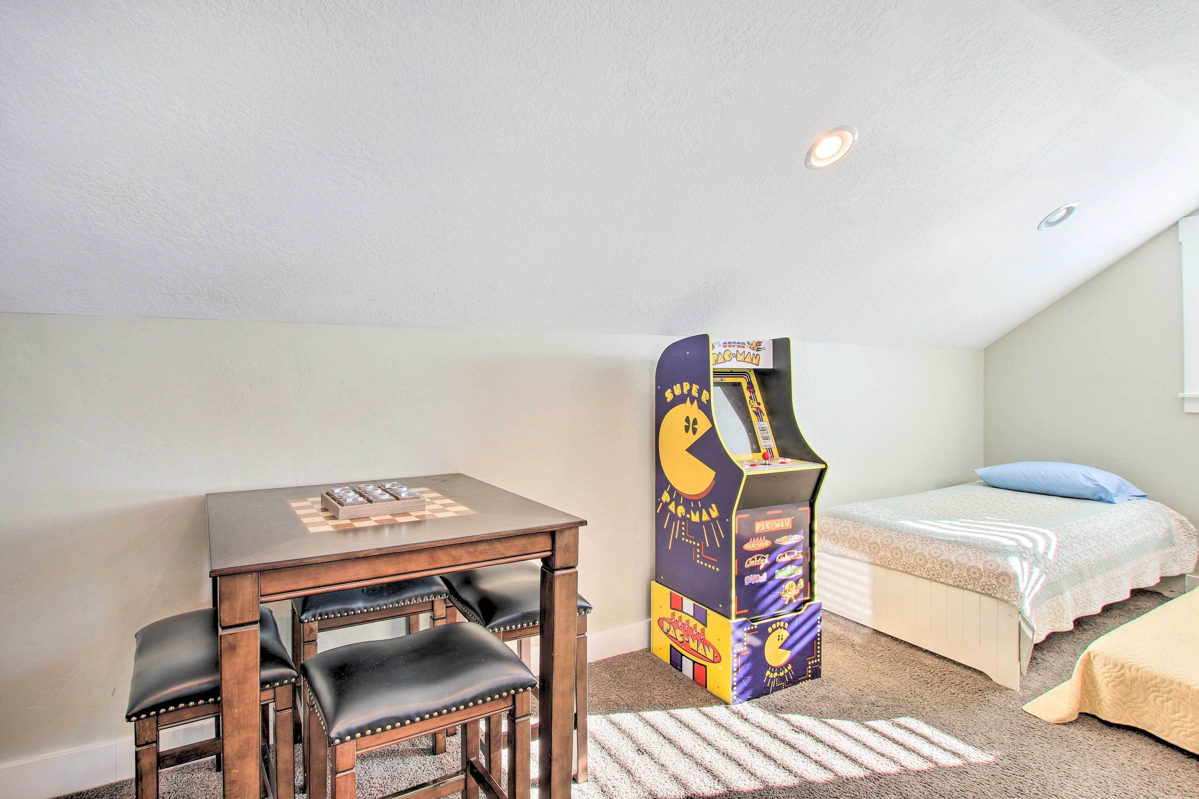 A room with a wooden table and chairs, a bed with a blue pillow, and a Pac-Man arcade machine.