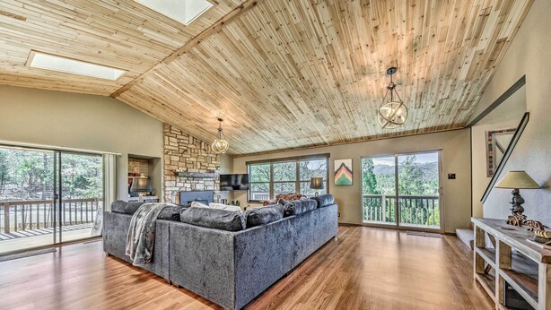 A spacious living area with a large gray sofa, wooden flooring, a stone fireplace, and large windows offering a view of the outdoors. - Ruidoso 'memories Mountain Retreat' w/ Hot Tub! (Ruidoso)