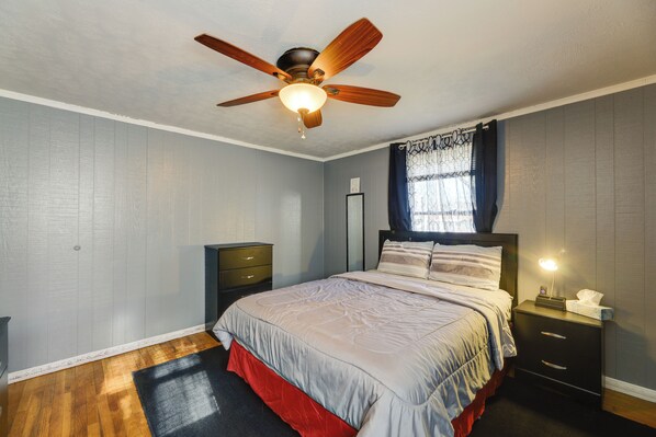 A bedroom with a bed, a nightstand, a ceiling fan, a window with curtains, and a wooden floor.