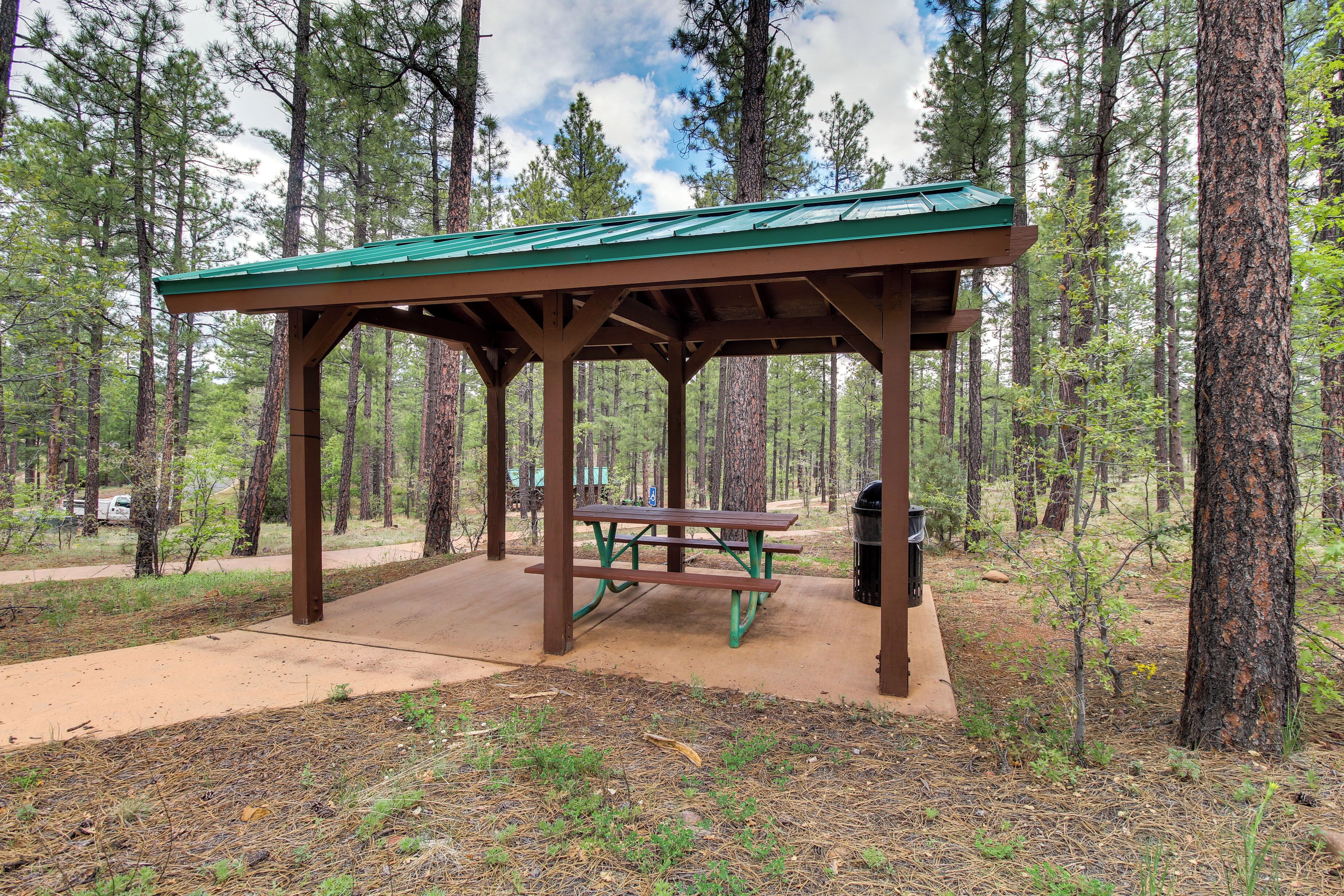 A covered picnic area with benches and a trash can in a forest setting.
