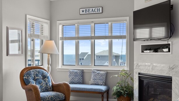 A living room with a wicker chair, a bench, a fireplace, and a large window with a view of the beach. - Avalon 507 3 Bedroom Condo by RedAwning (Ocean City)