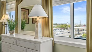 A hotel room with a white dresser, a mirror, a lamp, and a view of a cityscape through large windows. - Avalon 507 3 Bedroom Condo by RedAwning (Ocean City)