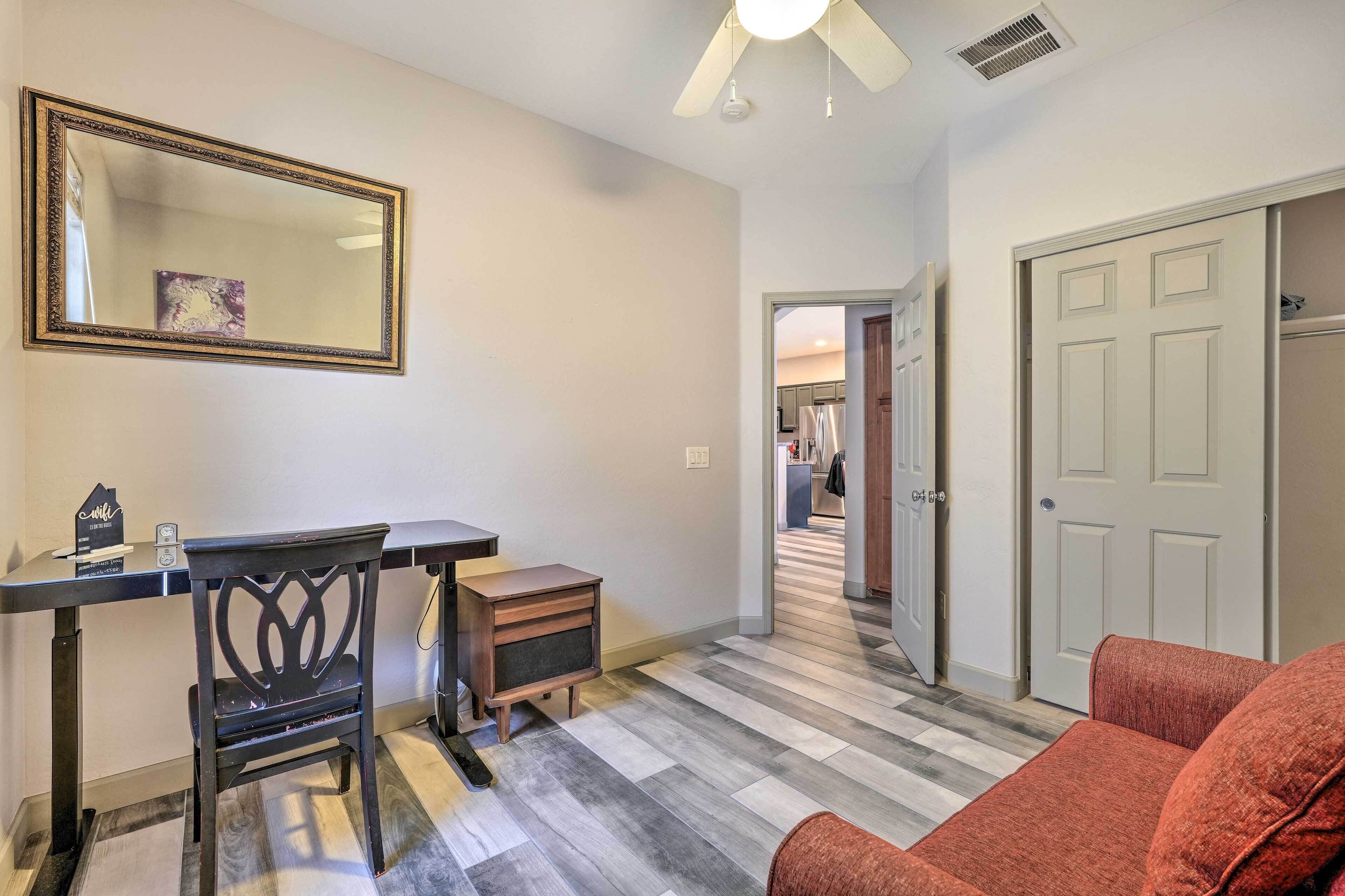 A room with a desk, chair, and a large mirror on the wall. There is a door on the right and a hallway leading to another room.