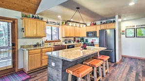 A modern kitchen with wooden cabinets, a granite island with bar stools, a stainless steel refrigerator, and a microwave.