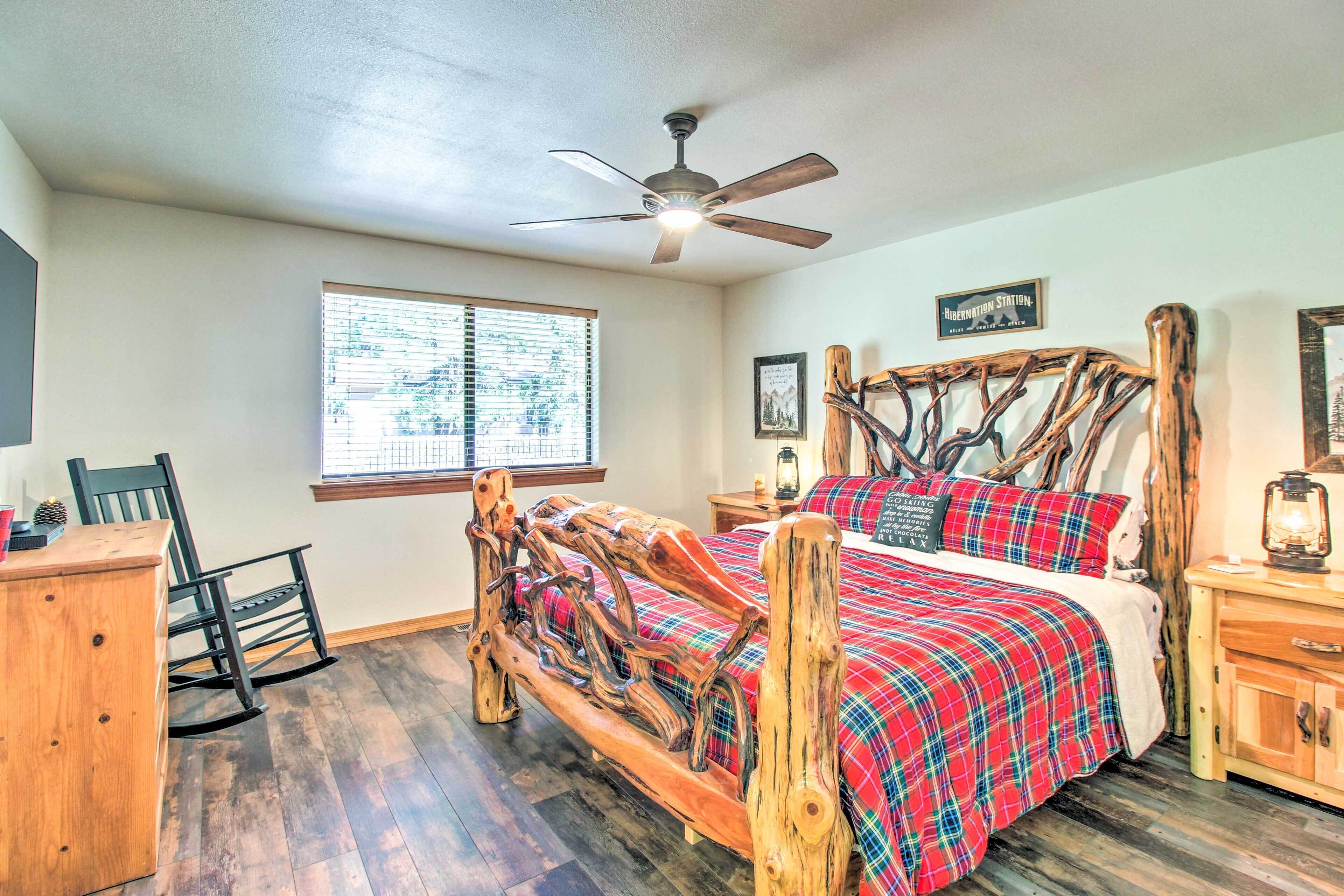 A bedroom with a wooden bed frame, a red plaid bedspread, a ceiling fan, a window with blinds, and a wooden nightstand with a lantern.