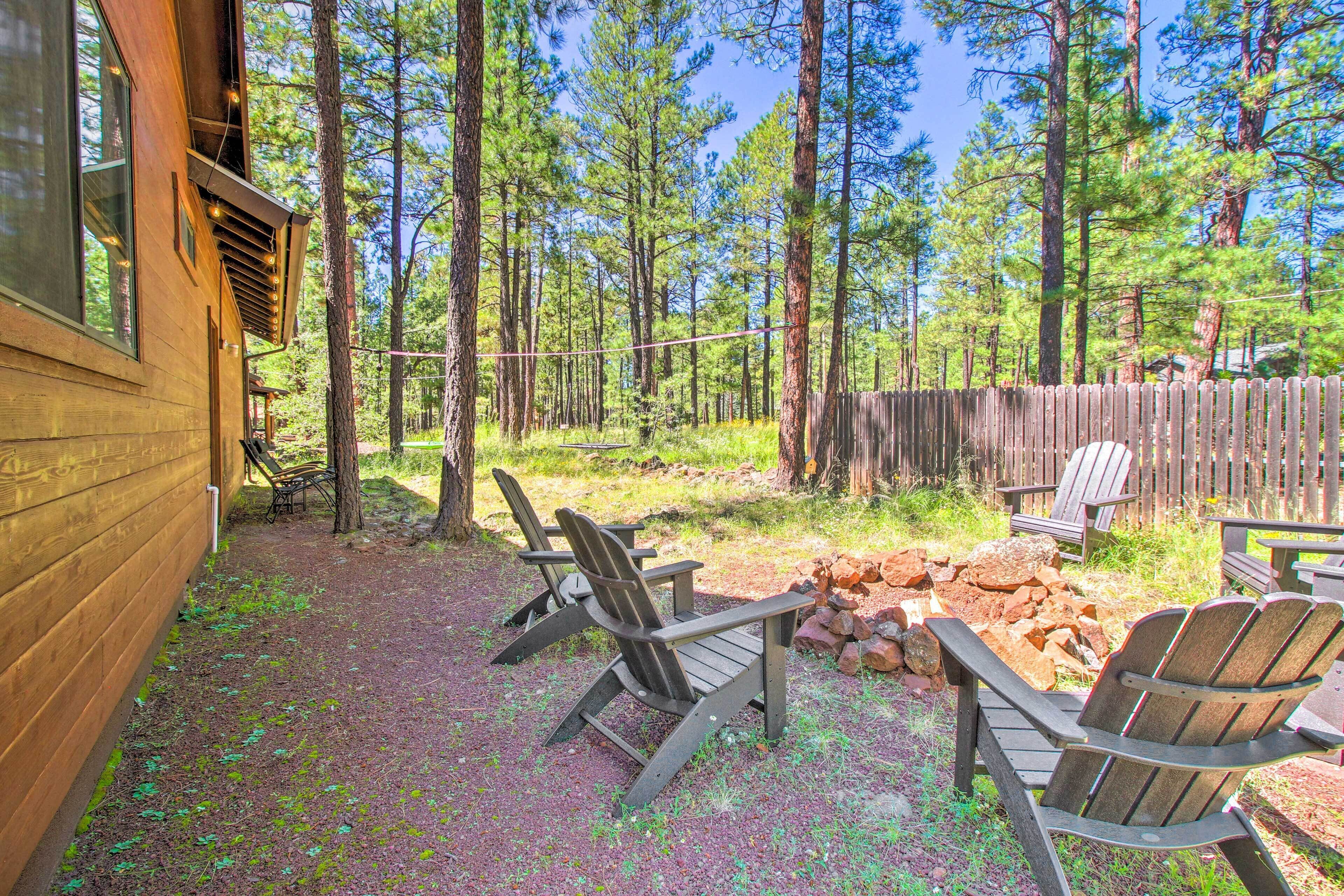 A wooden cabin with a deck, surrounded by a forest. There are Adirondack chairs and a fire pit.