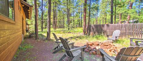 A wooden cabin with a deck, surrounded by a forest. There are Adirondack chairs and a fire pit.