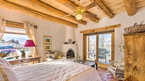 A bedroom with a large bed, wooden ceiling beams, a ceiling fan, a fireplace, and a view of the outdoors. - Casita Vacation Rental Near Taos w/ Patio! (Taos)