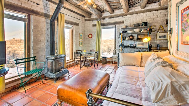 A rustic room with a brick wall, wooden beams, a large window, a green bench, a vintage stove, a dining table with chairs, and a bed with white bedding. - Casita Vacation Rental Near Taos w/ Patio! (Taos)
