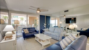 A modern living room with a blue sofa, striped armchair, and a glass coffee table. There is a ceiling fan, a TV mounted on the wall, and a large window with a view of palm trees. - Tranquil Redington Beach Condo w/ Beach Access (Redington Beach)