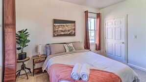 Un dormitorio con cama, mesita de noche, planta en maceta, un cuadro en la pared y una puerta.
